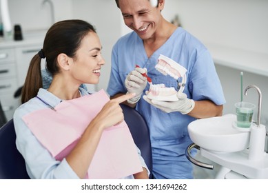 Portrait of beautiful young woman sitting in dental chair while stomatologist in sterile gloves holding false teeth and toothbrush. He is looking at brunette girl and smiling