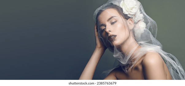 Portrait of a beautiful young woman with smooth skin. White flowers are woven into dark hair. White veil on the face. Fashionable makeup.Advertising concept for a wedding salon.