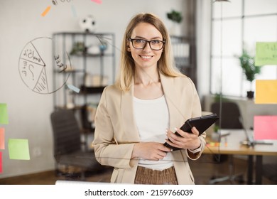 Portrait of beautiful young woman standing in front of glass board and smiling on camera. Female office worker in glasses and stylish suit using digital tablet in company. - Powered by Shutterstock