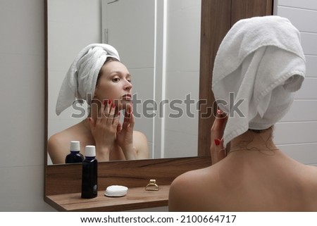 Portrait of a beautiful young woman standing in the bathroom and examining her face in the mirror, problematic acne-prone skin concept	
