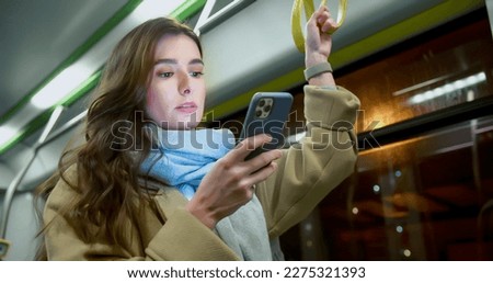 Portrait of beautiful young woman standing in public transport holding handrail texting on smartphone online.