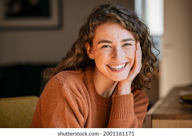 Portrait of beautiful young woman staying at home. Happy woman smiling while looking at camera with hand on cheek. Pretty laughing girl with curly hair and winter clothes relaxing in living room.