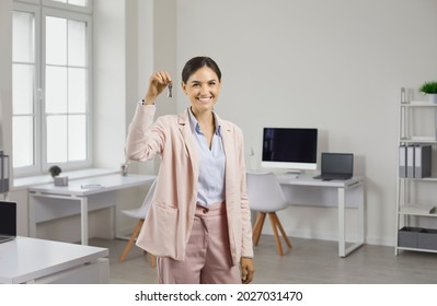 Portrait Of Beautiful Young Woman In Suit Standing In Office, Holding Keys And Smiling. Happy Realtor, Real Estate Agent, Mortgage Broker, Company Manager Giving Client Keys To New House Or Apartment