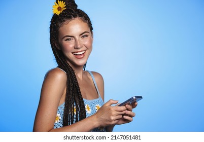 Portrait of beautiful young woman with summer tan, flower in hair, holding smartphone and smiling, using mobile phone on vacation, standing over blue background