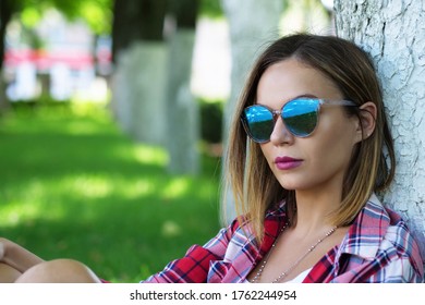 Portrait of beautiful young woman in sunglasses sitting under tree on green grass at city park.