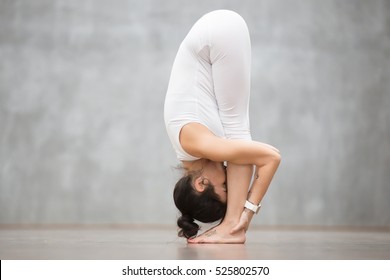 Portrait of beautiful young woman with tattoo on her foot meaning "Wild kitty" working out against grey wall, doing yoga or pilates exercise. Uttanasana, head to knees pose. Full length photo