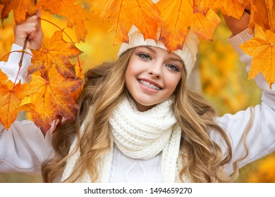 Portrait of beautiful young woman walking outdoors in autumn. Girl with autumn yellow leaf in knitted hat and sweater.  Portrait of a girl with orthodontic appliance.