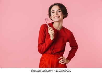Portrait of a beautiful young woman wearing red dress standing isolated over pink background, holding candy cane