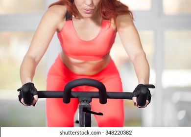 Portrait Of Beautiful Young Woman Wearing Red Bright Sportswear Exercising On Cycle In Sports Club, Warming Up During Sport Lesson In Class In Gym, Controlling Her Breath, Close-up