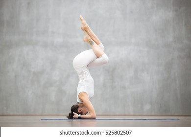 Portrait of beautiful young woman wearing white sportswear working out against grey wall, doing yoga or pilates exercise. Headstand, Garuda salamba sirsasana. Full length photo