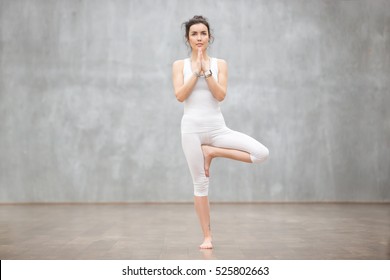 Portrait of beautiful young woman wearing white sportswear working out against grey wall, doing yoga or pilates exercise. Standing in Vrksasana, Tree pose. Full length