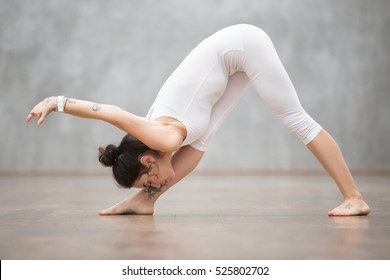 Portrait of beautiful young woman wearing white sportswear working out against grey wall, doing yoga or pilates exercise. Standing in Parsvottanasana, Pyramid, One Sided Fold pose. Full length