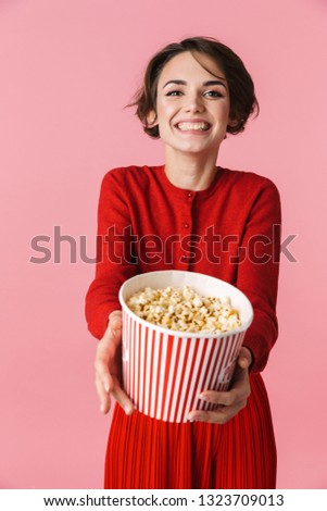 Portrait of a beautiful young woman wearing red dress standing isolated over pink background, eating popcorn