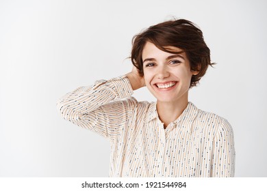 Portrait of beautiful young woman with white smile, standing in blouse and touching short hair, looking at camera, standing over white background. - Shutterstock ID 1921544984