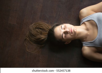 Portrait Of Beautiful Young Woman Working Out At Home, Doing Yoga Exercise On Wooden Floor, Lying In Shavasana (Corpse Or Dead Body Posture), Resting After Practice, Meditating, Breathing. Top View