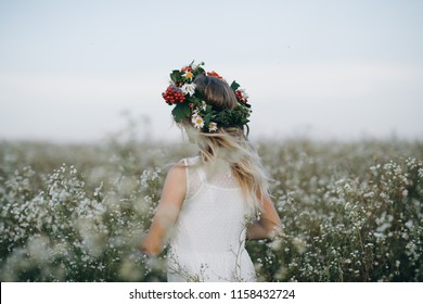 Portrait Of A Beauty Blonde Girl With Blue Eyes With A Wreath Of Flowers On Her Head Walking In Field With White Flowers