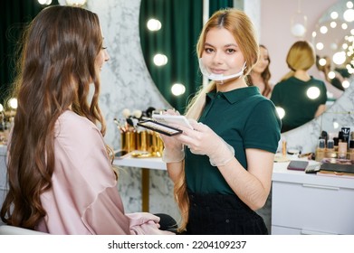 Portrait Of Beauty Specialist Holding Eyelashes Set While Standing Near Young Woman In Visage Studio. Female Makeup Artist In Face Mask Looking At Camera While Client Sitting On Chair In Beauty Salon.