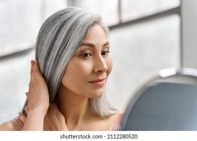 Portrait Of Beautyful Happy Middle Aged Mature Asian Woman, Senior Older 50s Lady Pampering Touching Gray Hair Looking At Herself At Mirror Indoors. Ads Of Lifting Anti Wrinkle Skin Hair Care Spa.