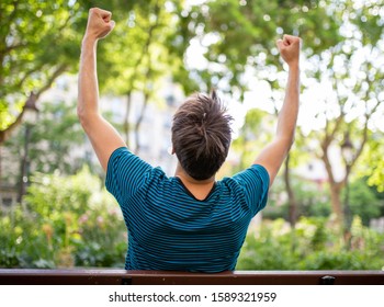 Portrait From Behind Of Young Man Sitting On Park Bench With Arms Raised