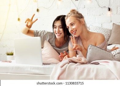 Portrait Of Best Friends Talking On Portable Computer Via Skype Lying And Laughing Together At Home In Bedroom. Happy Cute Women Showing Victory Symbols By Hands. Friendship Concept