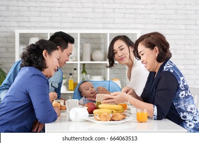 Portrait Of Big Asian Family At Breakfast, Enjoying Meal Feeding Cute Baby Boy Showing Him Cartoons On Smartphone  Happy Asian Family At Breakfast Table