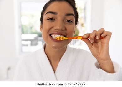 Portrait of biracial woman in bathrobe brushing teeth in sunny bathroom. Lifestyle, self care, hygiene and domestic life, unaltered. - Powered by Shutterstock