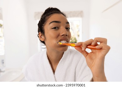 Portrait of biracial woman in bathrobe brushing teeth in sunny bathroom. Lifestyle, self care, hygiene and domestic life, unaltered. - Powered by Shutterstock