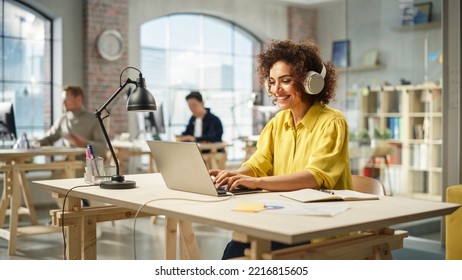 Portrait of Biracial Young Woman Smiling and Using Laptop in Bright Contemporary Office. Client Advisor Answering Customer Support Through Headphones and Typing. Team Working in the Background - Powered by Shutterstock