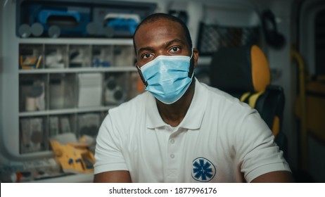 Portrait Of A Black African American EMS Paramedic Looks At Camera While Wearing A Safety Face Mask In Ambulance Vehicle. Emergency Medical Technician Outside The Hospital. Covid-19 Concept.