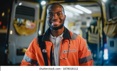 Portrait Of A Black African American EMS Paramedic Proudly Standing In Front Of Camera In High Visibility Medical Orange Uniform And Smiling. Successful Emergency Medical Technician Or Doctor At Work.