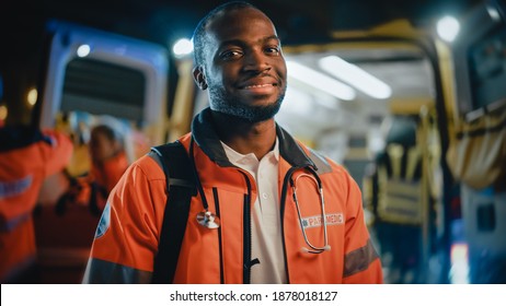 Portrait Of A Black African American EMS Paramedic Proudly Standing In Front Of Camera In High Visibility Medical Orange Uniform And Smiling. Successful Emergency Medical Technician Or Doctor At Work.