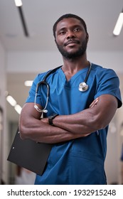 Portrait Of Black African Nurse Or Doctor In Blue Uniform Posing In Hospital Aisle With Arms Folded, With Stethoscope. American Medicine Concept