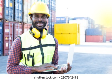 Portrait Of Black American African Warehouse Staff Wearing Yellow Hard Hat, Construction Vest And Ear Protector Standing With Happy Smile And Checking List Of Container Boxes For Shipping Logistic Stock.