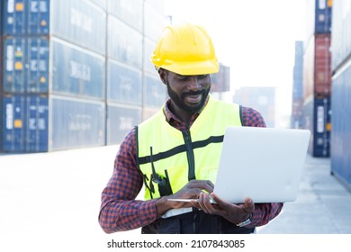 Portrait Of Black American African Warehouse Staff Wearing Yellow Hard Hat, Construction Vest And Ear Protector Standing With Happy Smile And Checking List Of Container Boxes For Shipping Logistic Stock.