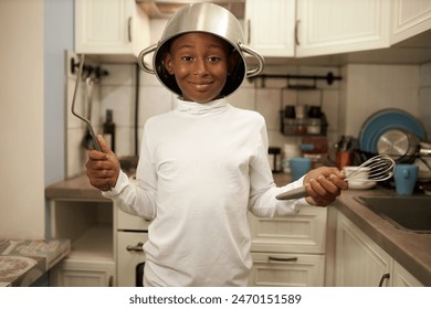 Portrait of black boy kid in blank mockup long-sleeve standing with cooking pan on head and whisk in hands, ready to help his mom to cook delicious dinner or bake cake for his birthday party