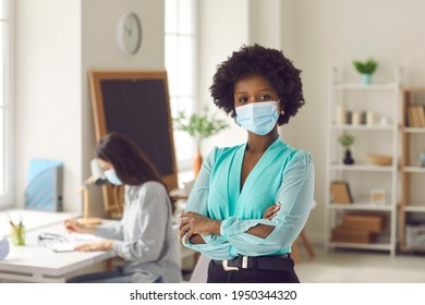 Portrait Of Black Businesswoman With Afro Hairstyle In Medical Face Mask Standing Arms Crossed In Office. Young Woman Returns To Work After Covid-19 Lockdown Ends. Coronavirus Pandemic Safety Concept