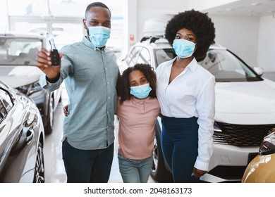 Portrait Of Black Family In Face Masks Showing Car Key, Buying New Vehicle In Auto Dealership During Covid Lockdown. Afro Parents With Cute Kid Purchasing Automobile At Showroom