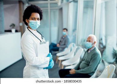 Portrait Of Black Female Doctor Wearing Protective Face Mask And Looking At Camera While Standing In A Waiting Room At The Hospital. There Are People In The Background. 