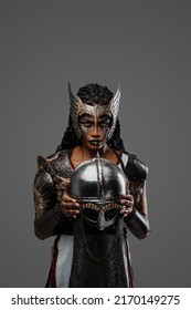 Portrait Of Black Female Warrior Dressed In Ancient Armor Holding Helmet Against Grey Background.