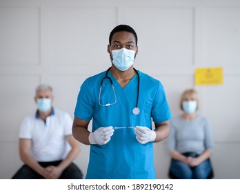 Portrait of black male doctor in protective mask and uniform holding syringe with coronavirus vaccine at hospital, patients waiting for covid-19 injection. Medical treatment concept