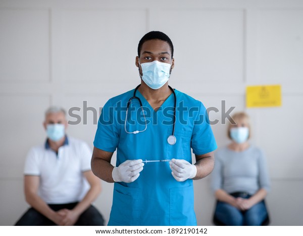 Portrait of black\
male doctor in protective mask and uniform holding syringe with\
coronavirus vaccine at hospital, patients waiting for covid-19\
injection. Medical treatment\
concept
