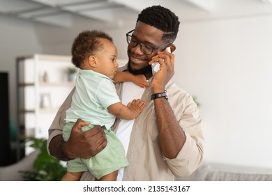 Portrait Of Black Millennial Man Holding Baby Toddler On Hands And Talking On Cell Phone, Spending Time With Little Son At Home. Smiling Father Working Remotely Making Call. Babysitting