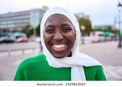 Portrait of black muslim woman very happy, smiling and looking at camera. An African student sincerely smiles while standing outdoors. The girl is wearing a white hijab and green t-shirt - Powered by Shutterstock