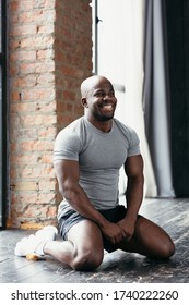 Portrait of a black pilates trainer in a gray t-shirt sitting on his knees on the floor in the gym and laughing.