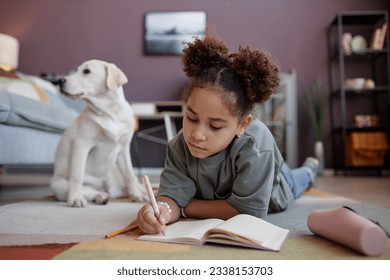 Portrait of black preteen girl writing in diary lying on floor at home with dog in background, copy space