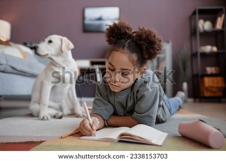 Portrait of black preteen girl writing in diary lying on floor at home with dog in background, copy space
