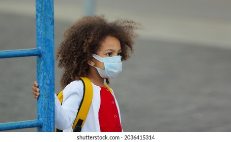 Portrait Of A Black Schoolgirl In A Medical Disposable Mask With A Backpack After School On The Playground. The Child Looks Into The Distance