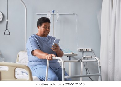 Portrait of black senior woman using mobile phone while sitting alone in hospital room - Powered by Shutterstock