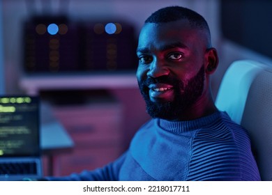 Portrait Of Black Software Engineer Looking At Camera While Sitting At Workplace In Neon Lights