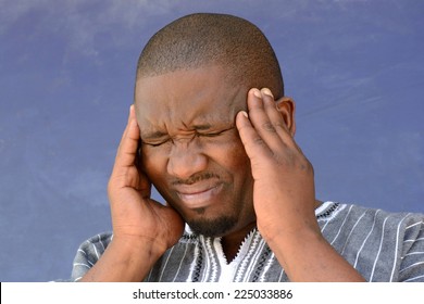 Portrait Of A Black South African Young Man With A Headache And A Sick Facial Expression Holding His Head With His Hands. Image On A Blurry Blue Dirty Wall Background.